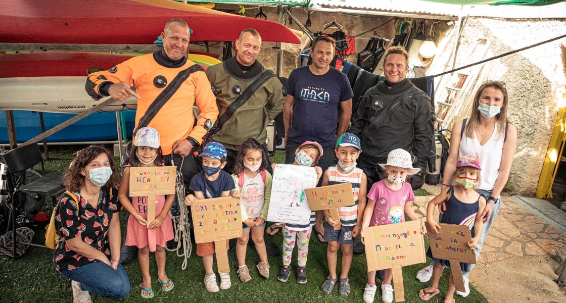 The Healthy Seas team with a group of children from the local community on Ithaca. The children all have masks on and are holding signs with messages written in Greek on them.