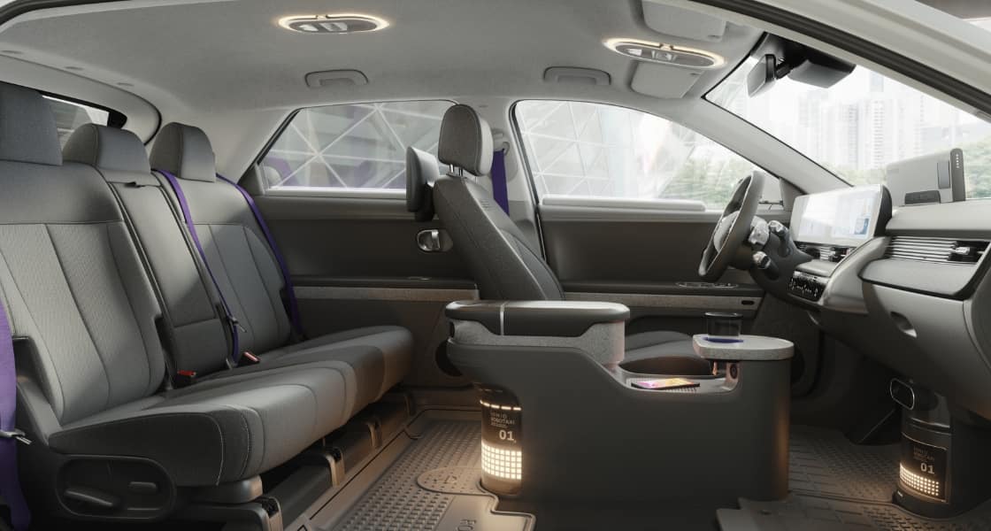The spacious interior inside a Hyundai’s IONIQ 5-based robotaxi. There are 2 rear seats and one front seat with purple seatbelts facing a steering wheel and dashboard.