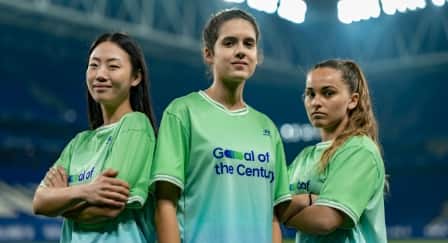 Three women standing in the middle of a stadium wearing green jerseys which say “Goal of the Century” on the front. 
