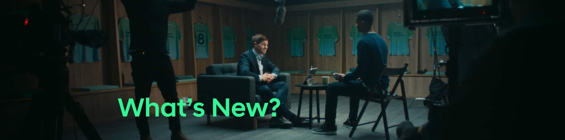Steven Gerrard sitting in the changing room surrounded by Team Century jerseys, being interviewed. ‘What’s New?’ is written in green across the image.